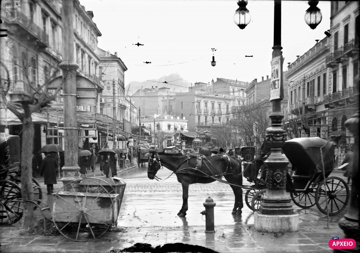 Βροχερή μέρα στην Αθήνα του 1927. Η οδός Πανεπιστημίου από την πλατεία Ομονοίας, μόνιππα αμάξια, αυτοκίνητα και πεζοί με ομπρέλες (φωτ. Αρχείο ΕΡΤ). 